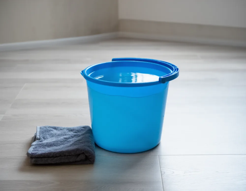 A bucket of clean water and a folded microfibre cloth on light laminate flooring